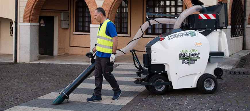 Street cleaning worker in yellow safety vest operating a compact street sweeping machine on a cobblestone plaza in front of a historic building with arched doorway. The white mechanical sweeper has FOLLOW ME branding visible. Urban municipal cleaning service in action.